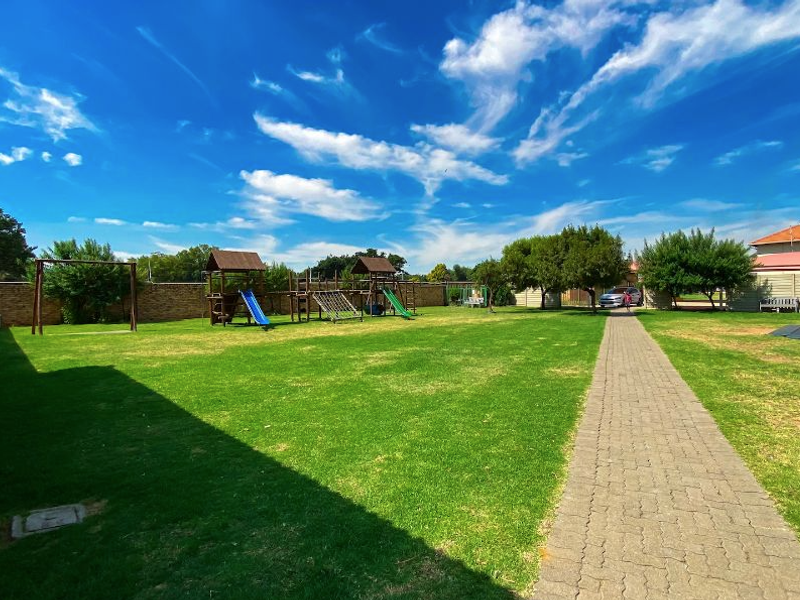 A well-maintained communal garden area featuring a paved walkway, expansive lawn, and fixed playground equipment within a secure boundary wall.