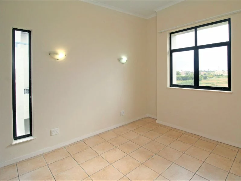 A bright, empty bedroom featuring neutral tiled flooring, wall-mounted lighting, and large windows with views of the surrounding area.