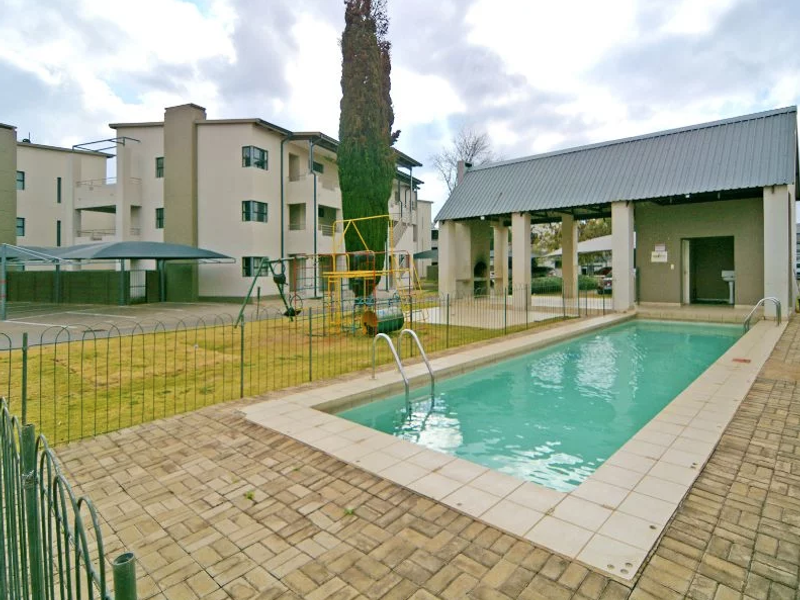Communal pool area featuring a paved deck, covered entertainment space with built-in braai, and surrounding metal fencing within an apartment complex.