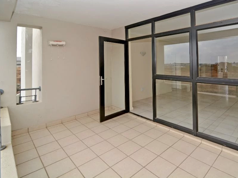 A spacious, empty balcony featuring tiled flooring, a black aluminum-framed glass door, and a large glass partition, offering a clean and functional outdoor space.