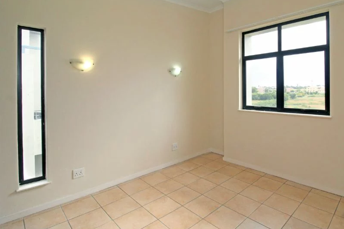 A bright, empty bedroom featuring neutral tiled flooring, wall-mounted lighting, and large windows with views of the surrounding area.