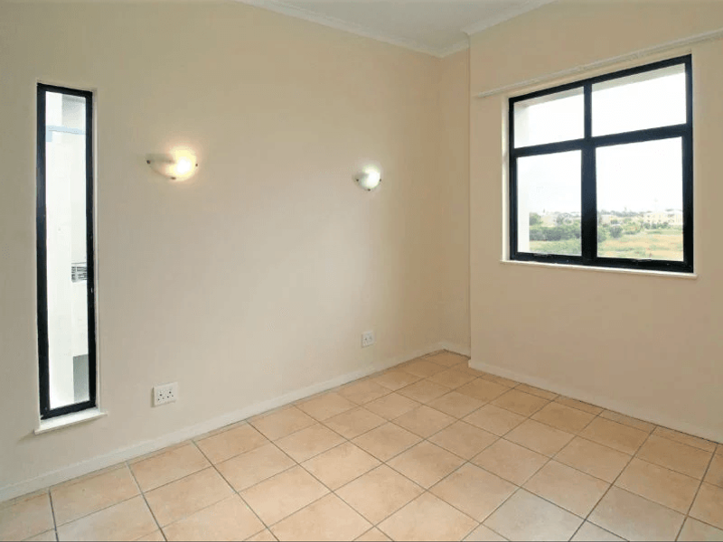 An empty bedroom featuring neutral tiled flooring, aluminium-framed windows, and wall-mounted lighting fixtures, offering a clean and versatile space.