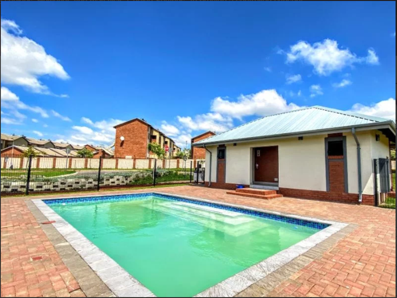 A communal swimming pool area featuring brick paving, a metal-roofed clubhouse structure, and perimeter fencing within a residential apartment complex.
