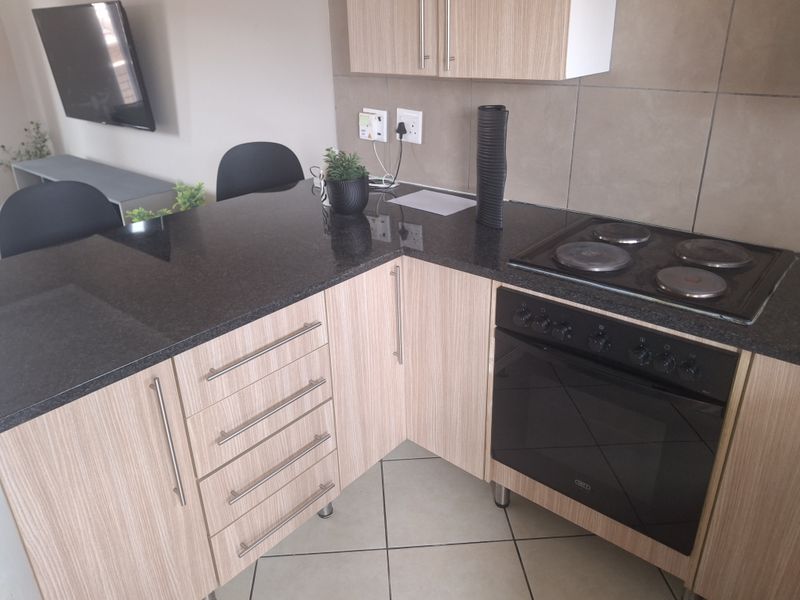 A compact kitchen featuring light wood-finish cabinetry, dark granite countertops, a built-in electric oven and hob, and neutral tiled flooring.