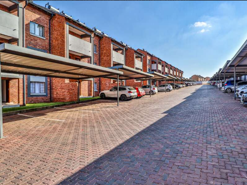 Exterior view of an apartment complex featuring a paved parking area with covered bays and brick building facades.