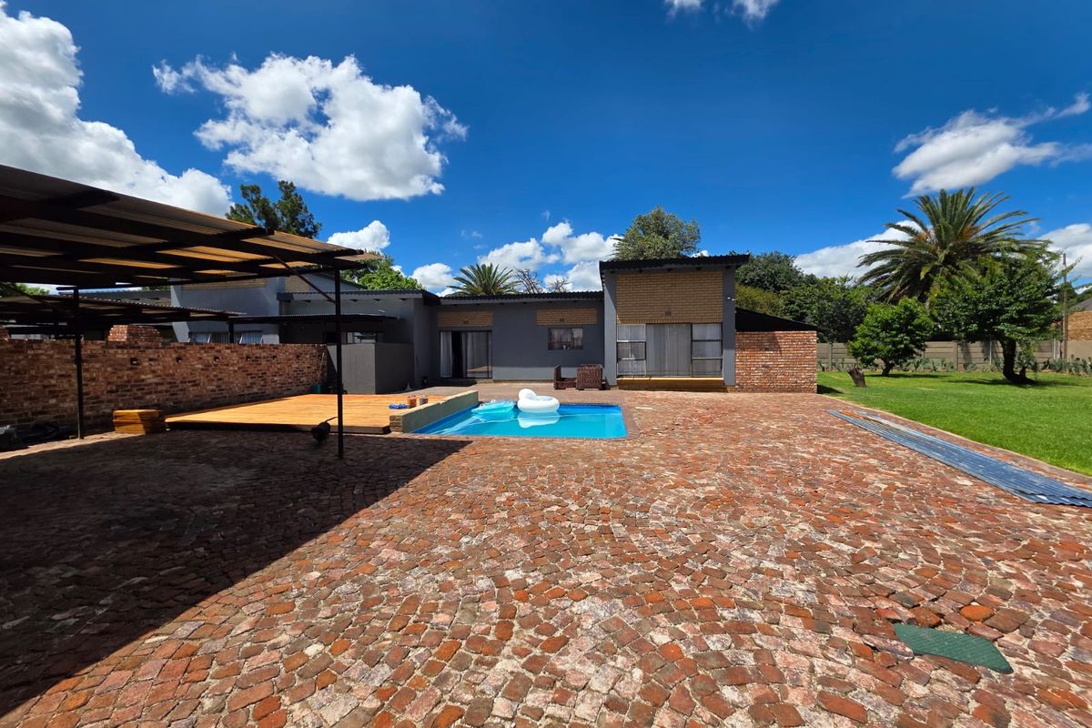 Spacious rear exterior featuring a swimming pool, wooden deck, covered patio area, and extensive brick-paved courtyard set within a large garden.