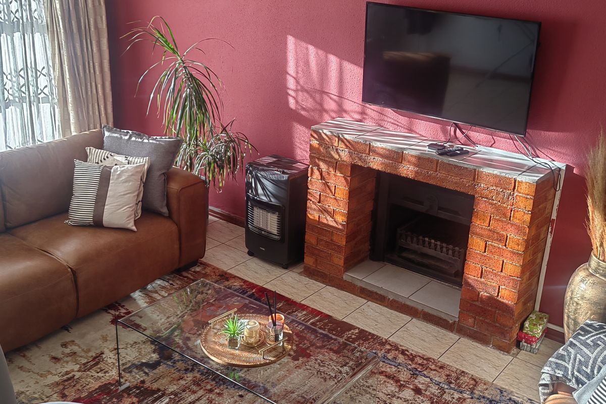 A cozy living room featuring a prominent brick fireplace, tiled flooring, and natural light entering through a window with a security gate.
