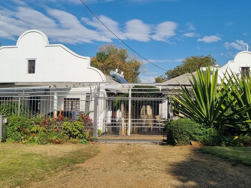 Front view of a property featuring distinctive Cape Dutch style gables, a secure metal gate, and established garden landscaping.