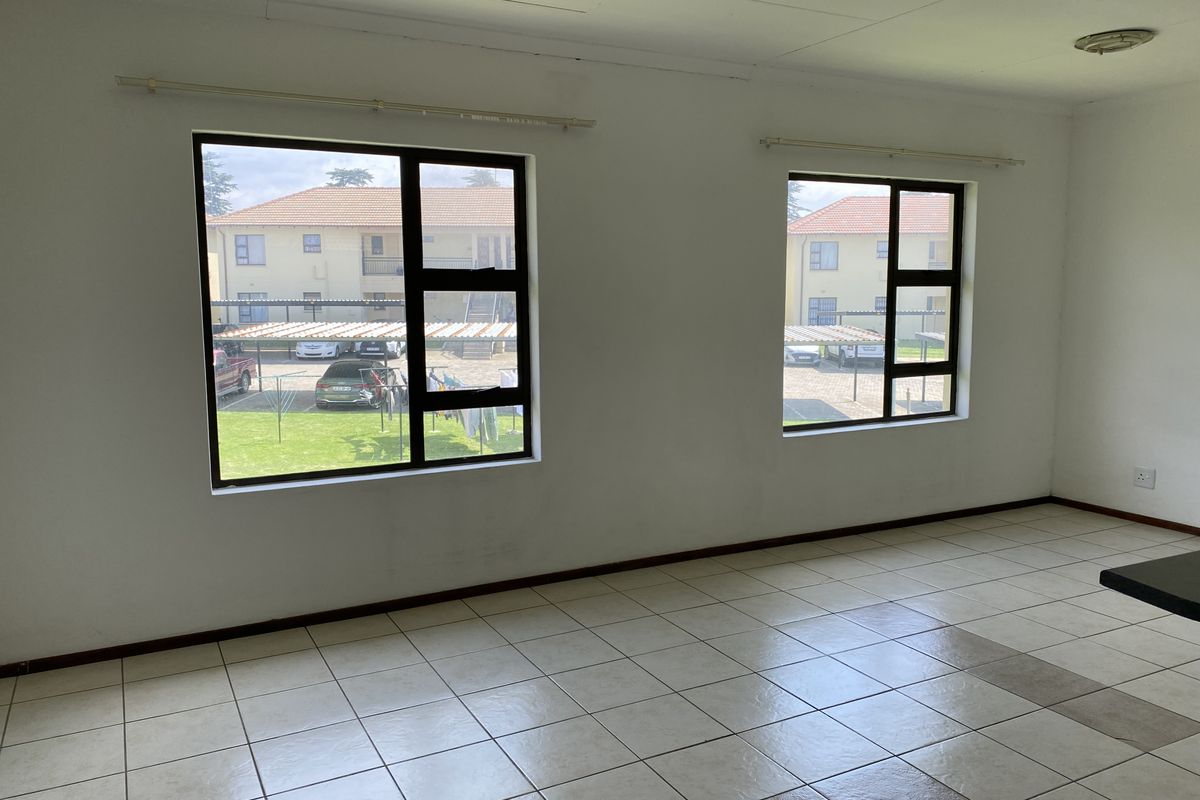 An empty, neutrally painted living area featuring tiled flooring and large aluminium-framed windows providing natural light and views of the complex exterior.