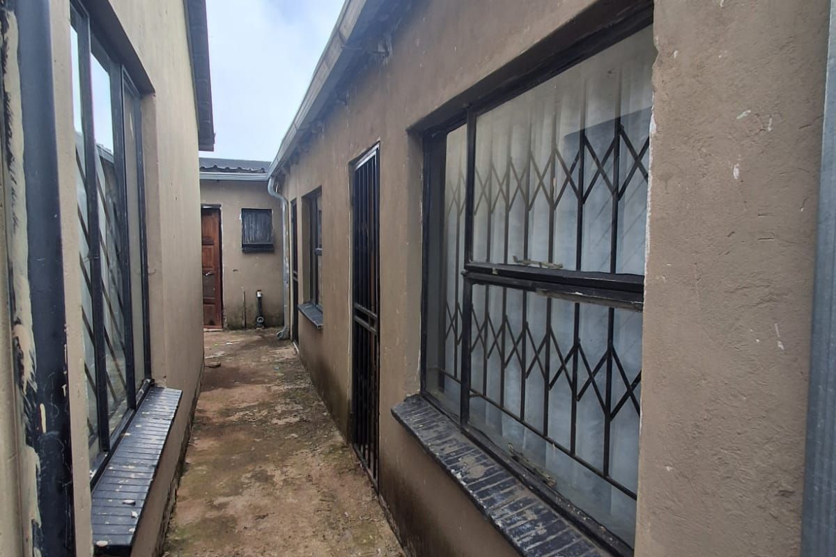 A narrow side passage between two building structures featuring concrete paving, security gates, and metal-framed windows with burglar bars.
