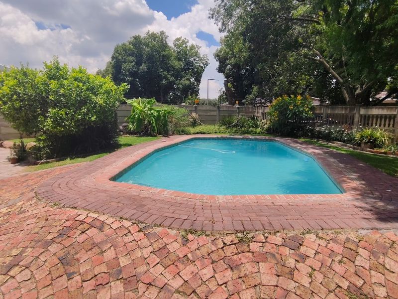 A private outdoor swimming pool area featuring a brick-paved surround, established garden landscaping, and perimeter boundary walling.