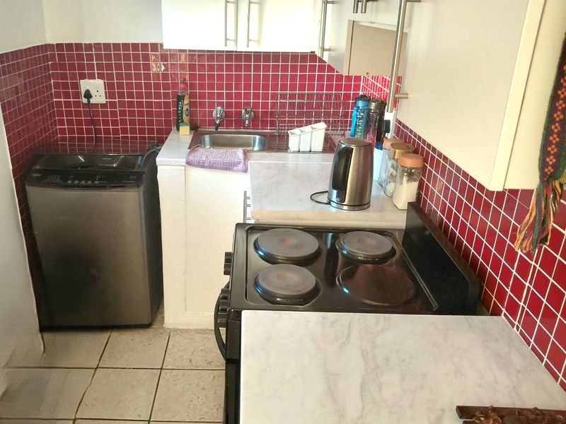 Compact kitchen featuring built-in cabinetry, a stainless steel sink, electric stove, and red tiled backsplash with tiled flooring.