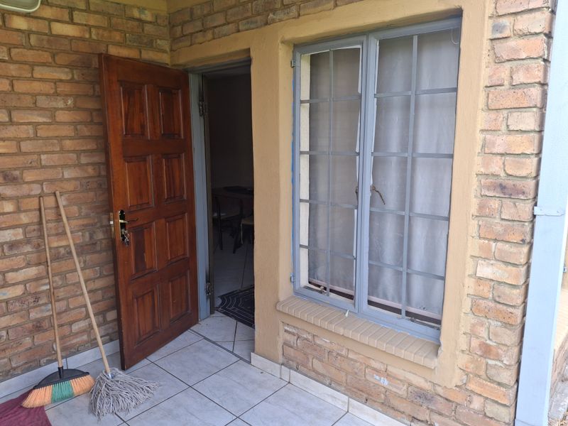A covered patio area featuring exposed brick walls, tiled flooring, and a wooden entrance door with an adjacent window protected by security bars.