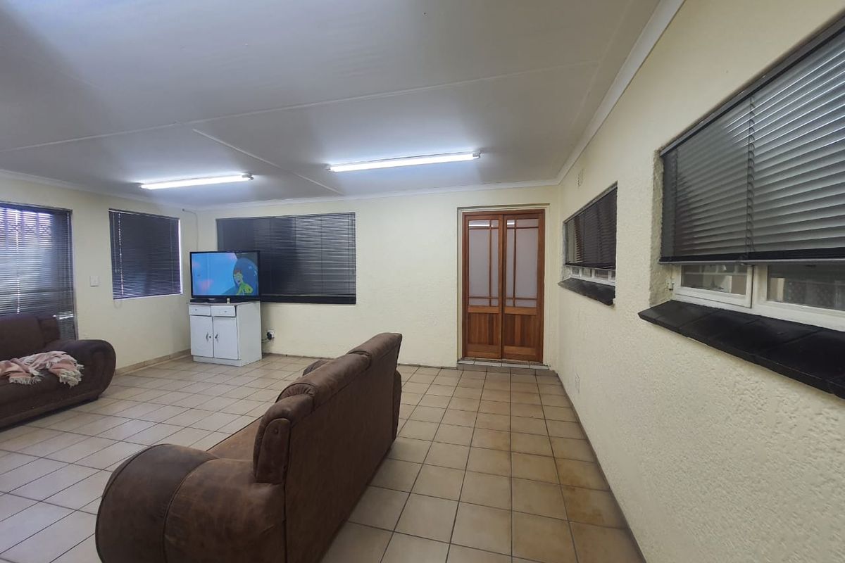 A spacious living room featuring tiled flooring, multiple windows with fitted blinds, and a wooden double door entrance.