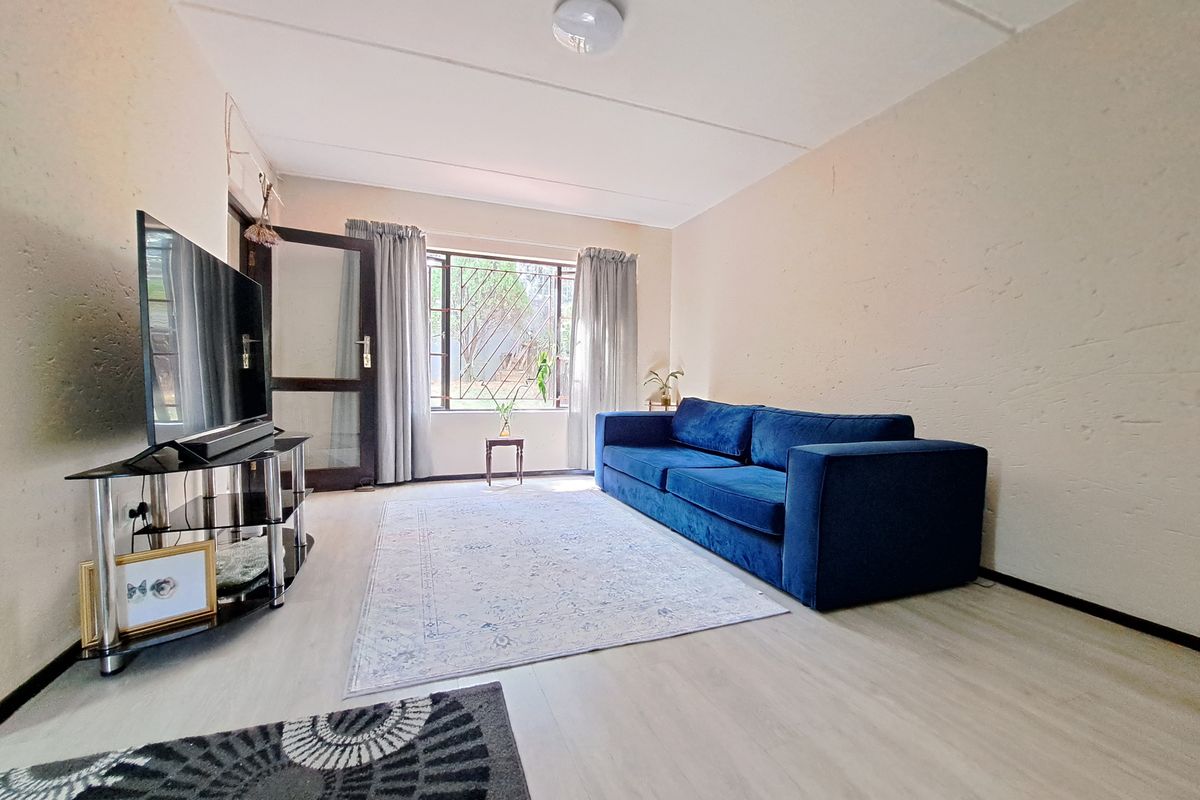 A living room featuring light-toned laminate flooring, textured walls, and a large window with a security gate, providing natural light to the space.
