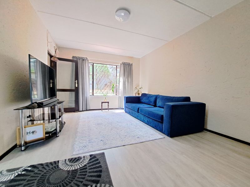 A living room featuring light-toned laminate flooring, textured walls, and a large window with a security gate, providing natural light to the space.