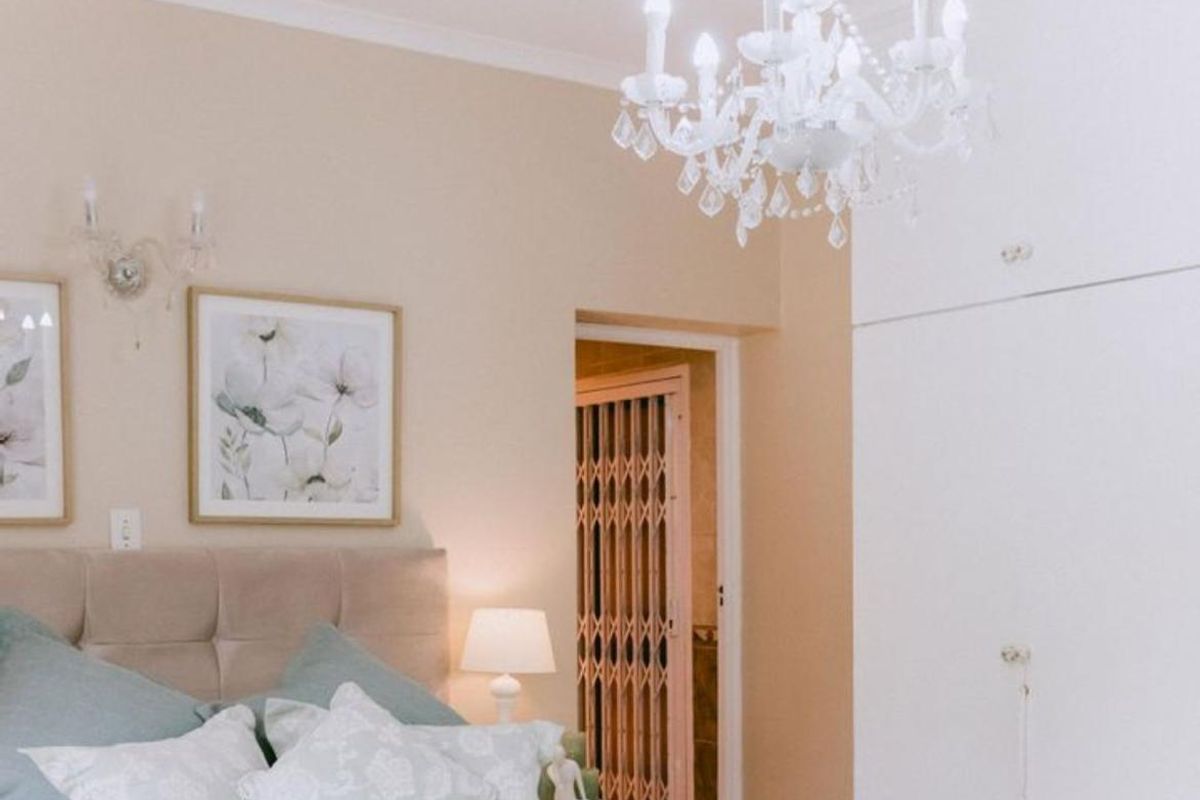 A well-maintained bedroom featuring built-in cupboards, a decorative chandelier, and an adjoining ensuite bathroom with a security gate.