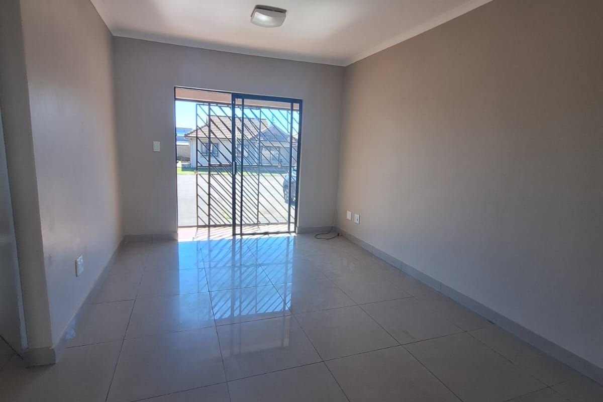 A bright, empty living room featuring neutral tiled flooring, smooth walls, and a sliding glass door with security gate leading to the exterior.