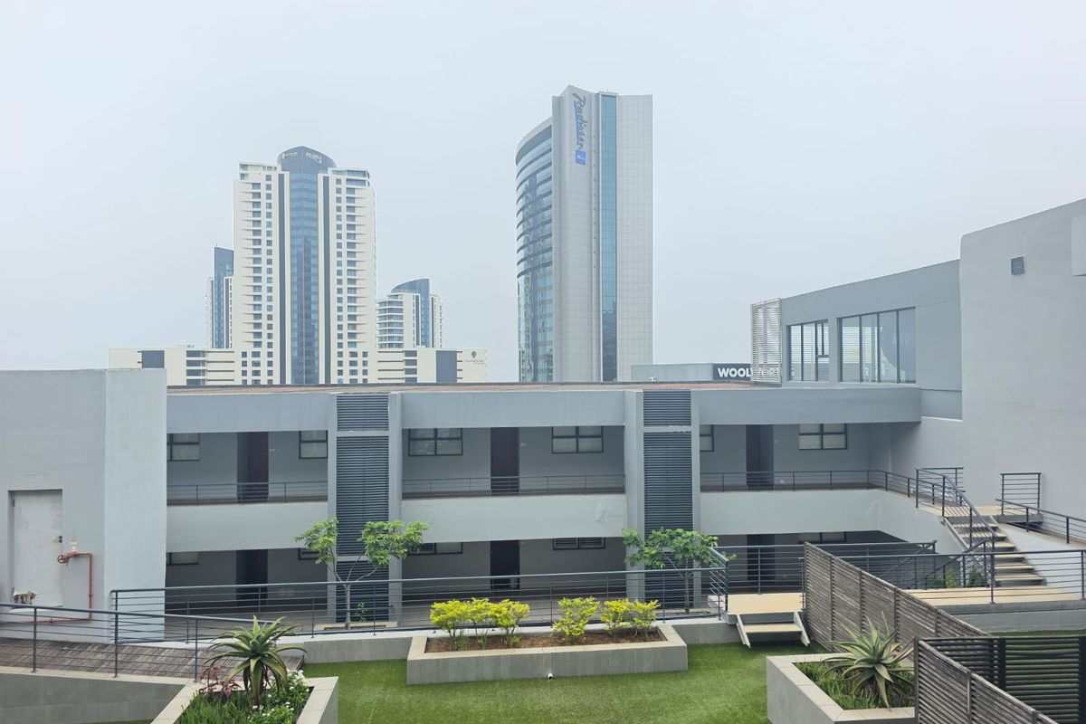 A view of the apartment complex exterior featuring landscaped garden beds, modern walkways, and a clear view of the surrounding city skyline.