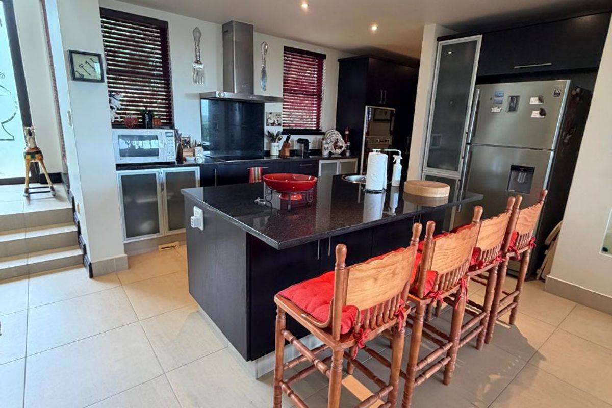 Modern kitchen featuring a central island with granite countertops, dark cabinetry, integrated appliances, and recessed ceiling lighting, flowing into an adjacent living area.