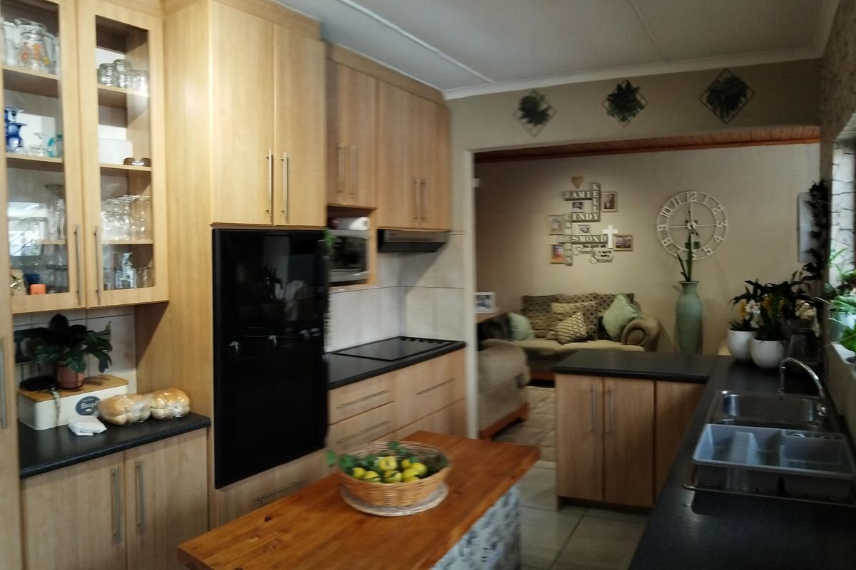 A functional kitchen featuring light wood cabinetry, dark countertops, a central island, and integrated appliances, with an open-plan flow into the adjacent living area.