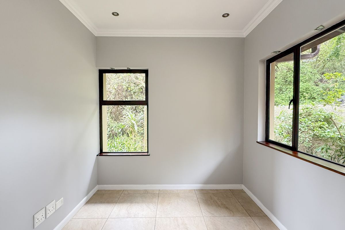 A bright, empty room featuring neutral wall paint, tiled flooring, recessed ceiling lighting, and dark-framed windows overlooking a garden.