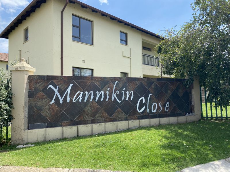 Front view of the Mannikin Close townhouse complex, featuring a stone-clad entrance wall, landscaped lawn, and multi-story residential building with aluminium window frames.