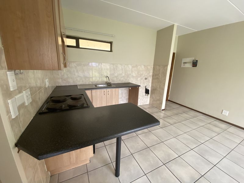 A functional kitchen featuring laminate cabinetry, an electric hob, stainless steel sink, and neutral ceramic tiling throughout the workspace and floor.