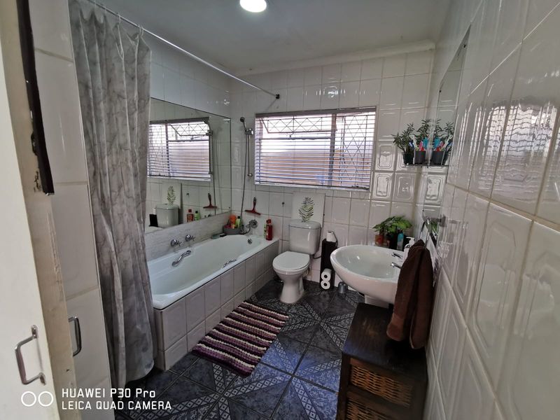 A functional bathroom featuring a built-in bathtub, pedestal washbasin, toilet, and fully tiled walls and floors with security bars on the window.