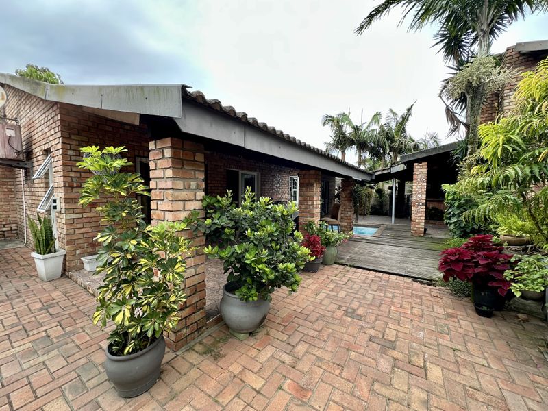 A covered outdoor patio area featuring exposed brick pillars, brick paving, and timber decking, leading towards a swimming pool in the rear garden.