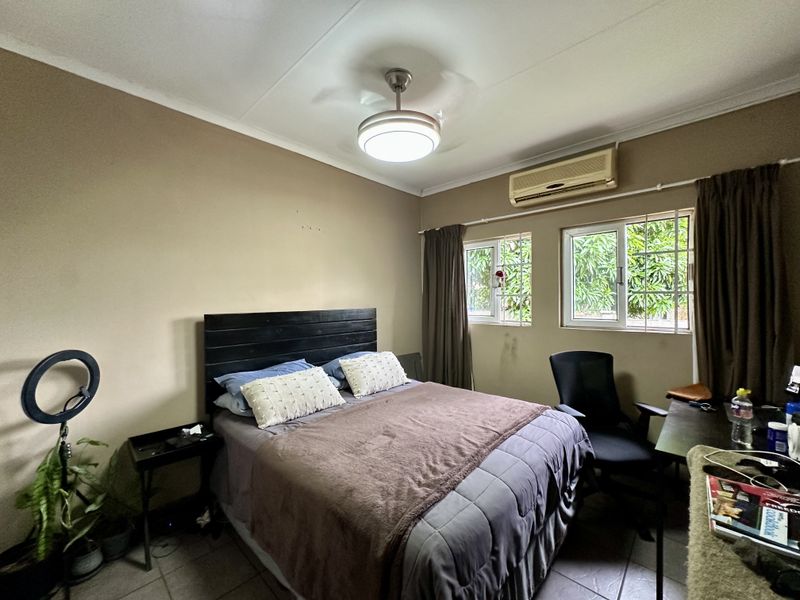 A functional bedroom featuring tiled flooring, a ceiling fan, and a wall-mounted split-system air conditioner with security bars on the windows.