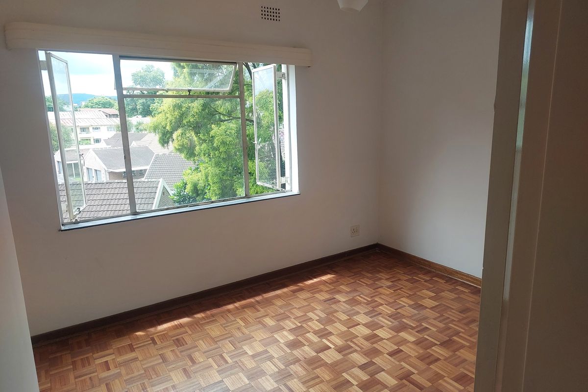 An empty bedroom featuring classic parquet flooring, neutral walls, and a large steel-framed window providing natural light and a view of the surrounding neighborhood.