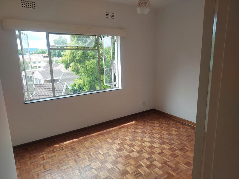 An empty bedroom featuring classic parquet flooring, neutral walls, and a large steel-framed window providing natural light and a view of the surrounding neighborhood.