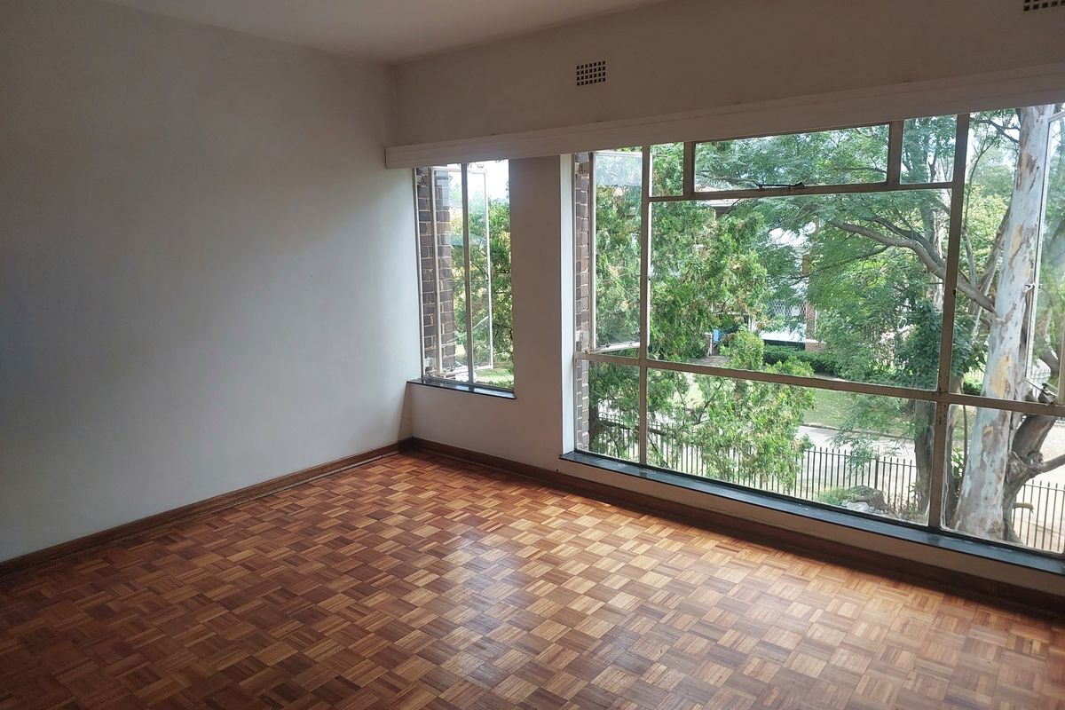 An empty living room featuring classic parquet flooring and large steel-framed windows that provide ample natural light and views of the surrounding greenery.