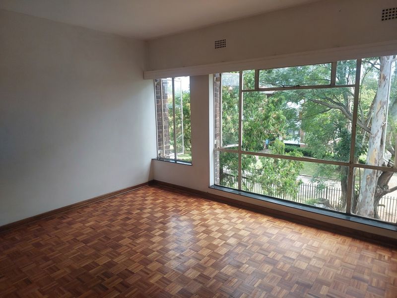 An empty living room featuring classic parquet flooring and large steel-framed windows that provide ample natural light and views of the surrounding greenery.