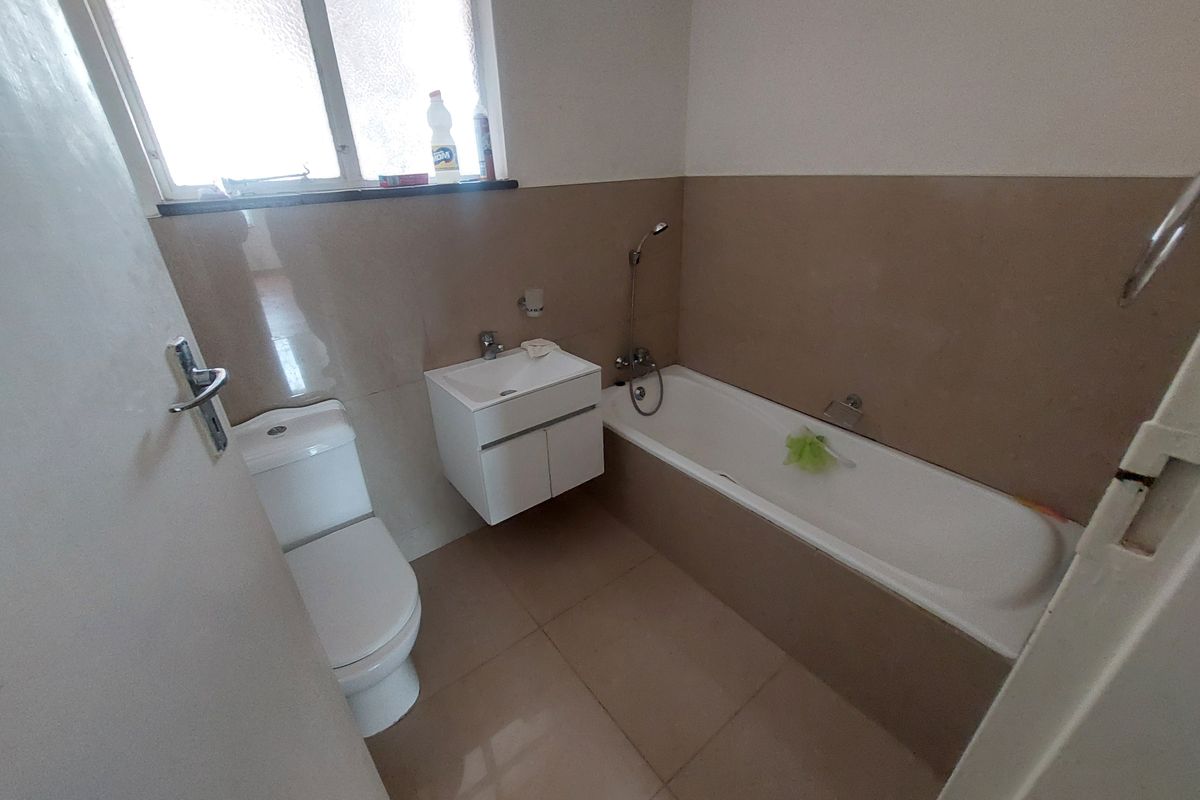 A functional bathroom featuring a bathtub with handheld shower, wall-mounted vanity, and toilet, finished with neutral wall and floor tiling.
