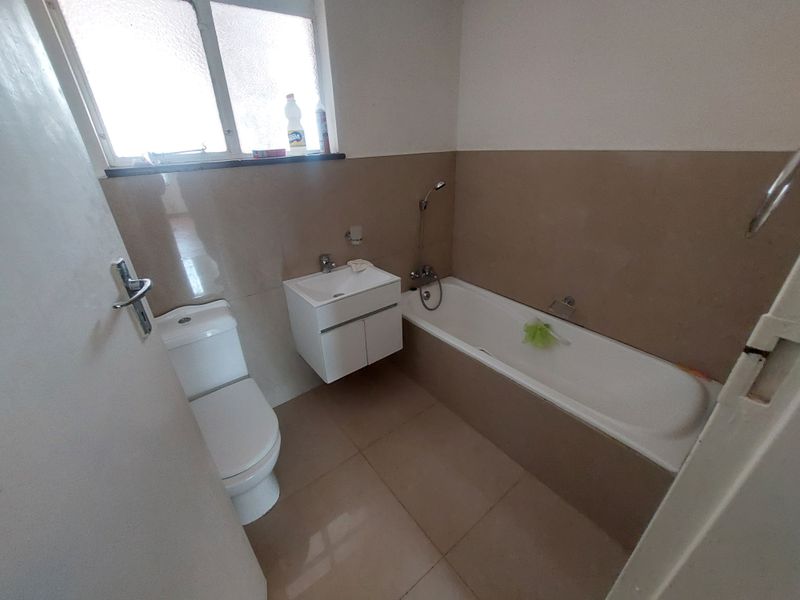 A functional bathroom featuring a bathtub with handheld shower, wall-mounted vanity, and toilet, finished with neutral wall and floor tiling.