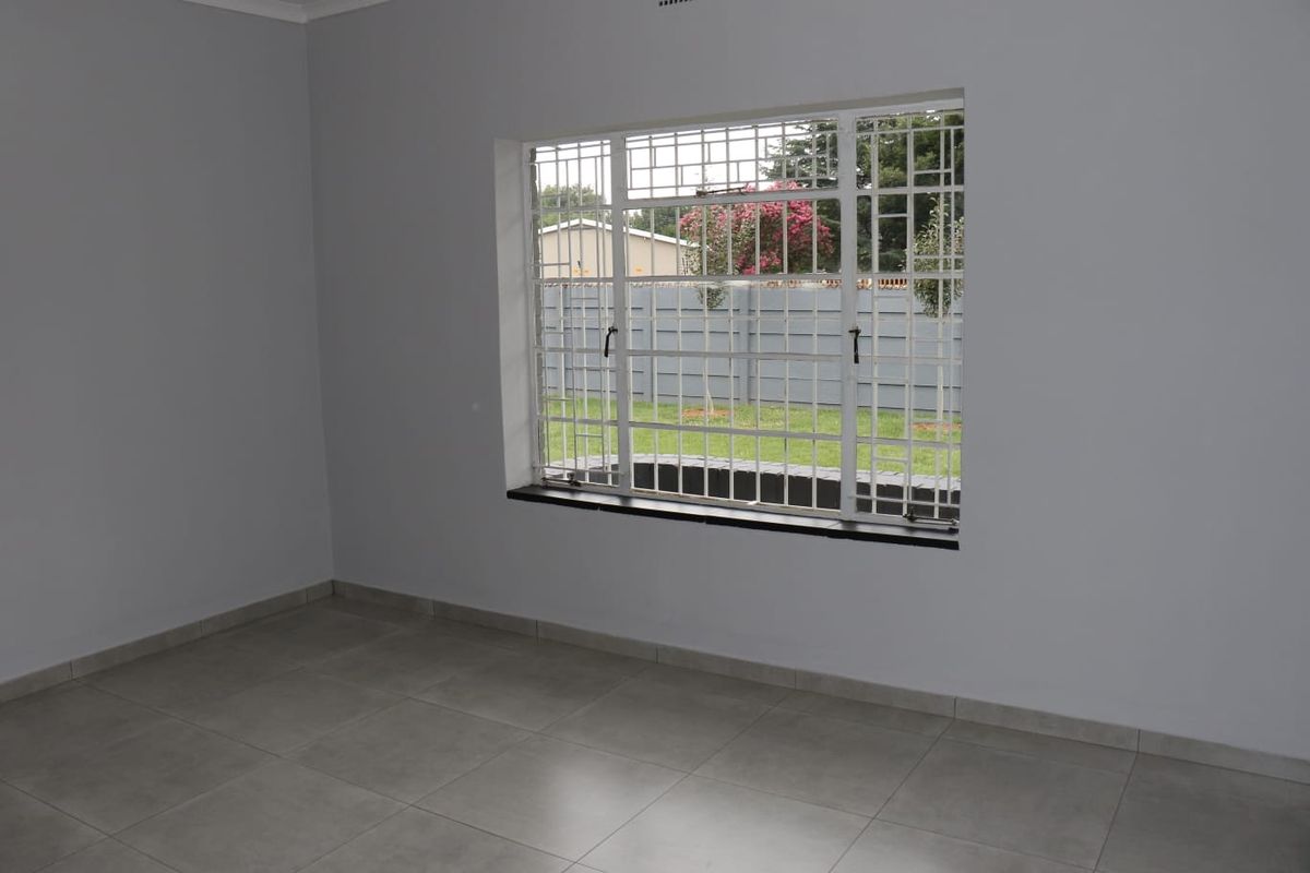 A bright, empty bedroom featuring neutral grey tiled flooring, freshly painted walls, and a large window fitted with security bars.