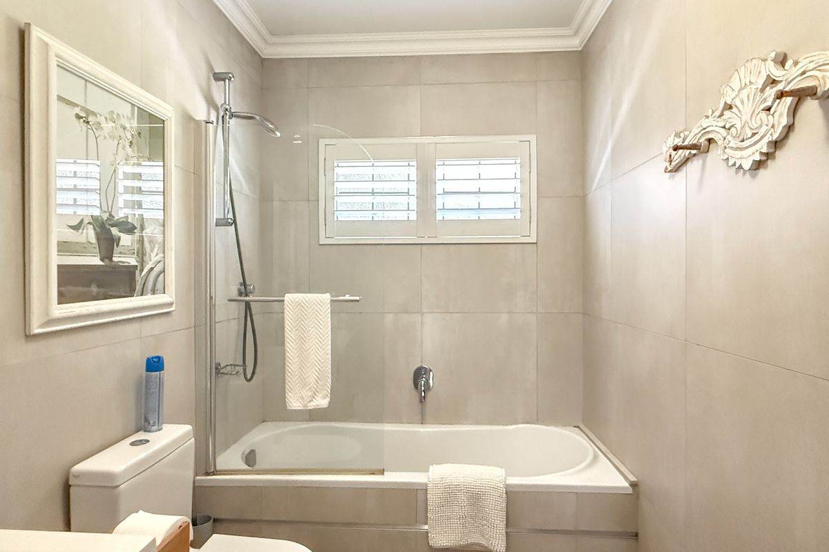 A clean, neutral-toned bathroom featuring a built-in bathtub with a glass shower screen, wall-mounted shower head, and plantation-style window shutters.