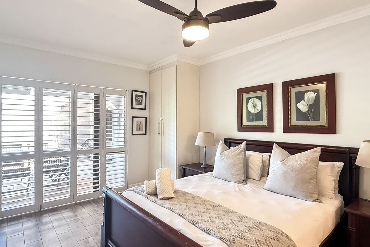 A well-maintained bedroom featuring tiled flooring, built-in cabinetry, a ceiling fan, and white plantation shutters opening onto a balcony.