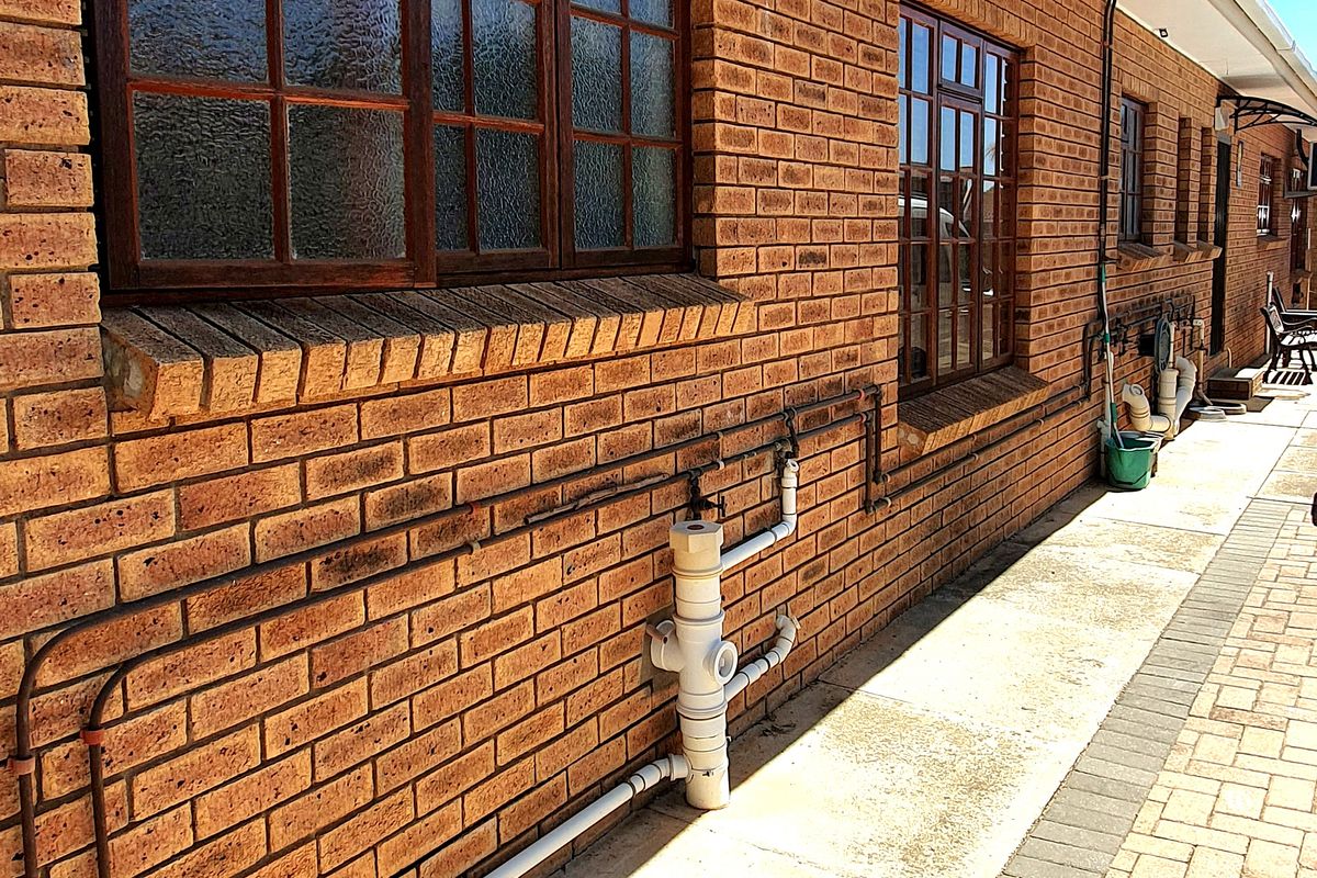 Side exterior view of the house featuring face-brick facade, timber-framed windows, and paved walkway with exposed exterior plumbing infrastructure.