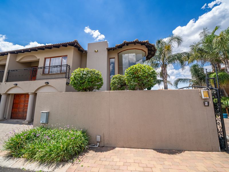 A well-maintained double-story residential facade featuring a balcony, arched architectural details, a tiled roof, and a secure perimeter wall with a driveway entrance.