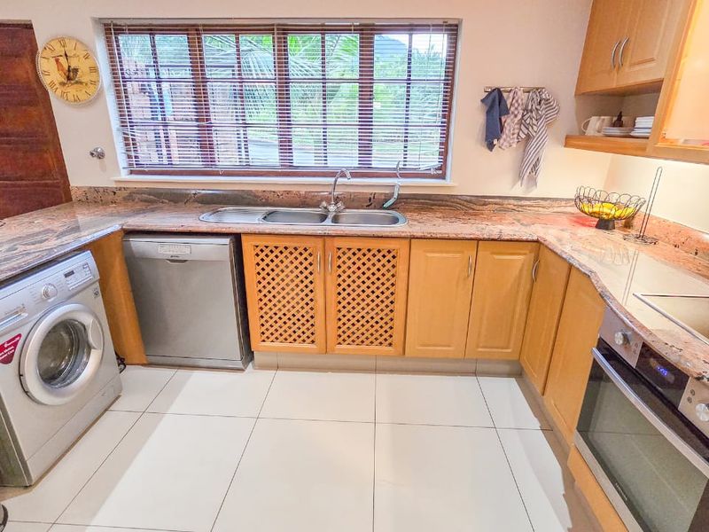 A functional kitchen featuring granite countertops, wooden cabinetry, built-in oven, electric hob, and tiled flooring with a double bowl sink under a window.