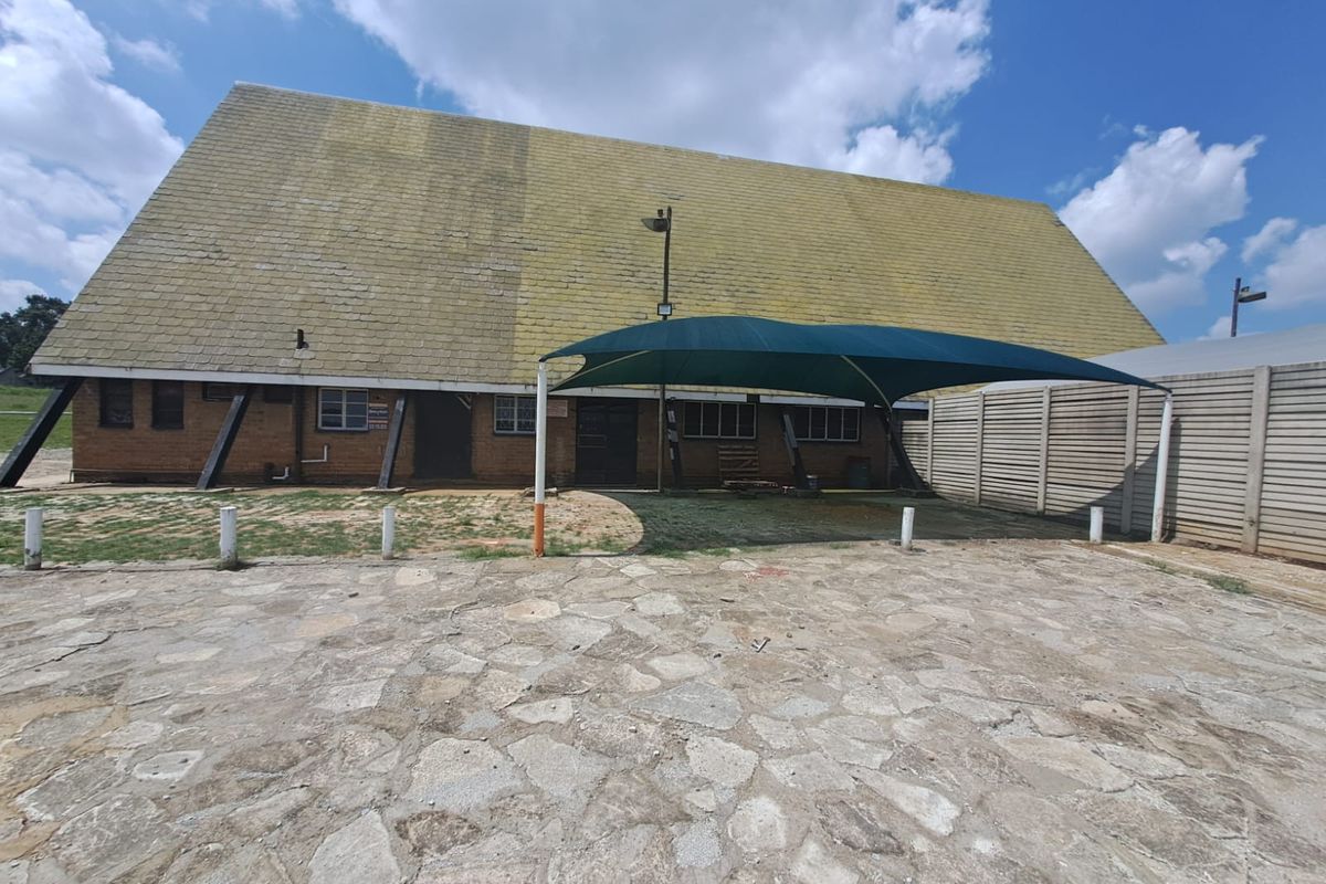 Rear exterior view of a farm building featuring a large pitched roof, a shade sail structure, and stone paved courtyard area.