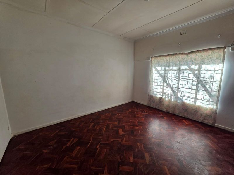 An empty bedroom featuring traditional parquet flooring, white walls, and a window fitted with security bars.