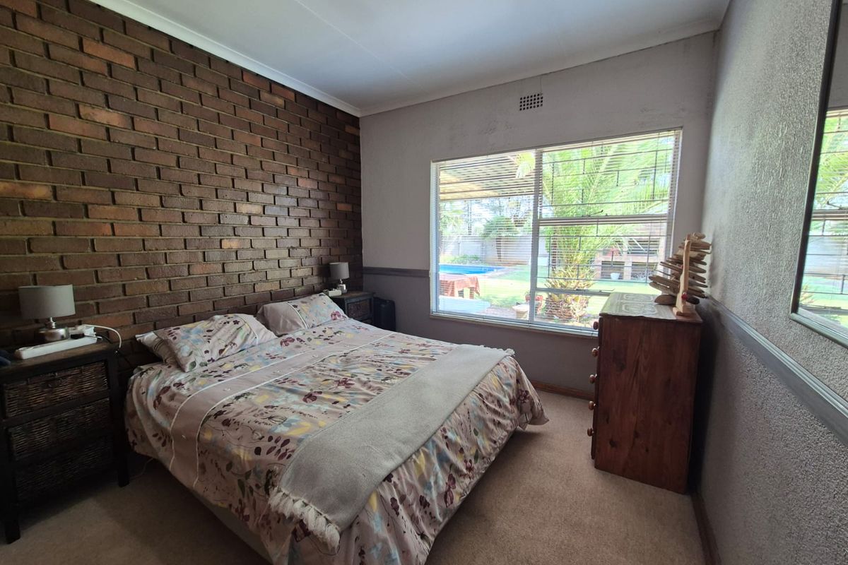 A bedroom featuring a distinctive exposed brick feature wall, carpeted flooring, and a large window overlooking the garden and pool area.