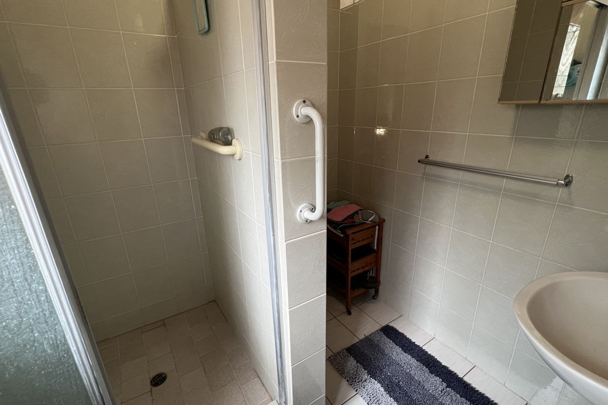 A functional bathroom featuring a walk-in shower enclosure with safety grab rails, a wall-mounted basin, and neutral wall tiling throughout.