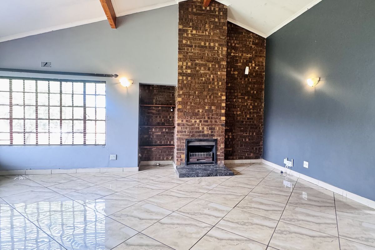 Spacious living room featuring a vaulted ceiling with exposed timber beams, large grid-style windows, and a prominent floor-to-ceiling brick fireplace.