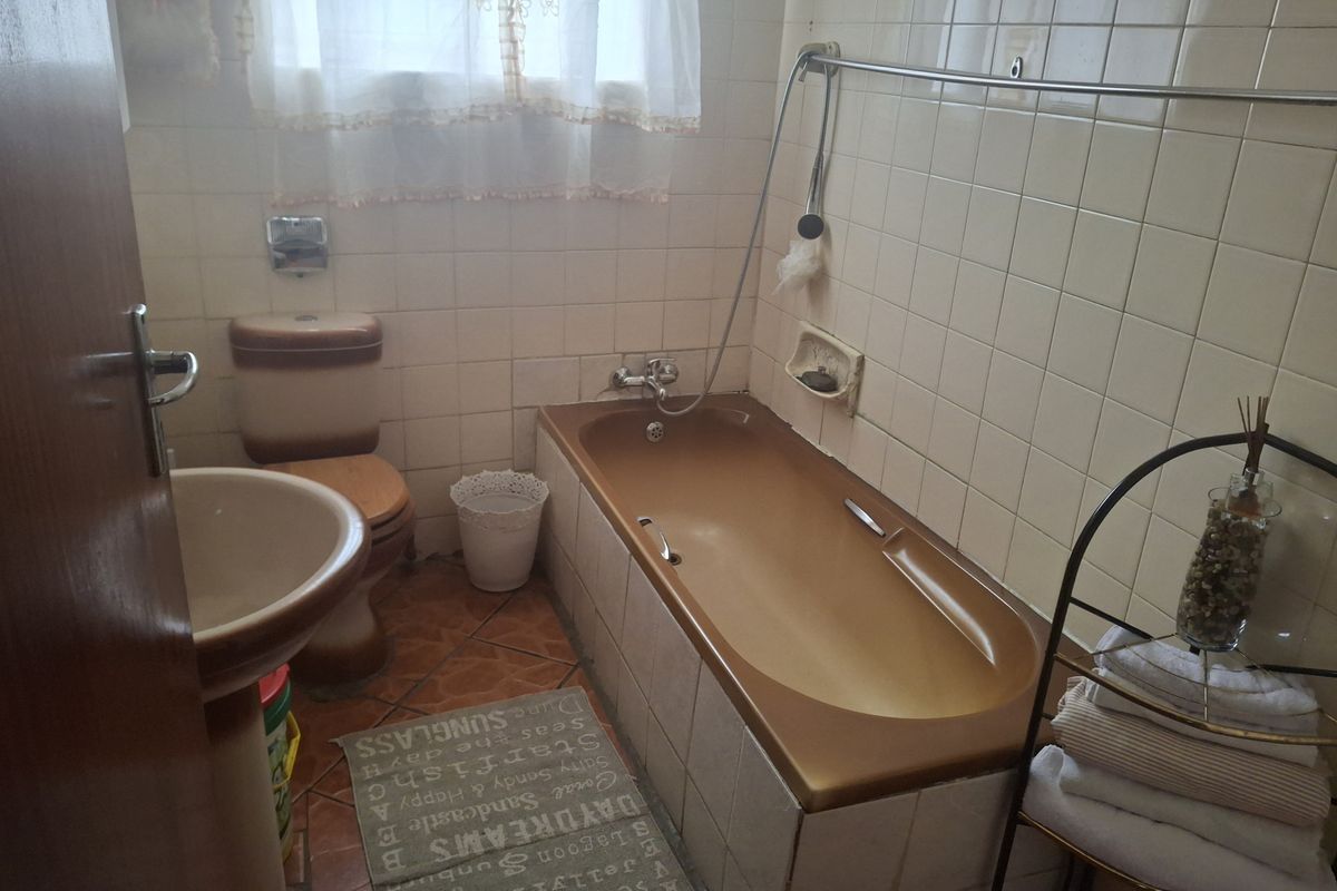 A dated bathroom featuring a pedestal basin, a built-in bathtub with wall-mounted shower mixer, and original ceramic wall and floor tiling.