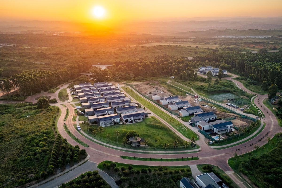 An aerial view of a modern residential estate featuring paved roads, landscaped surroundings, and a tennis court at sunset.